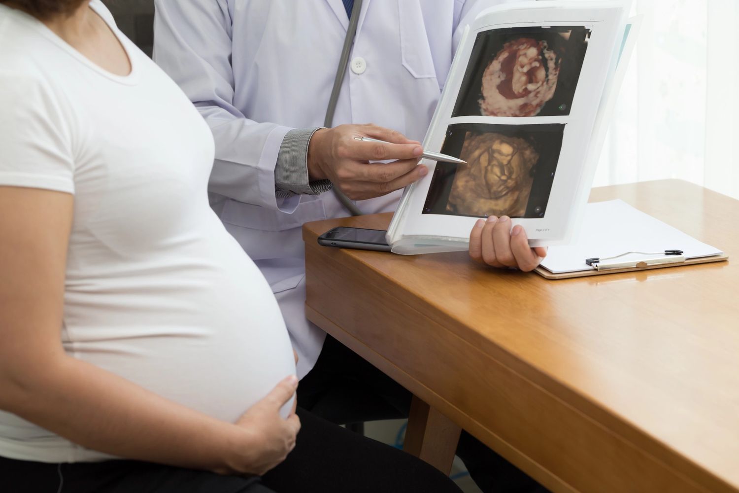 A doctor in a white clinical coat points to 4D ultrasound images in a patient file during a prenatal consultation with a pregnant woman in Houston.