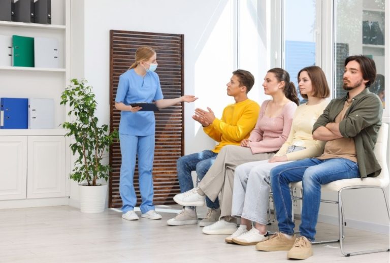 A nurse in blue scrubs and a mask addresses four patients sitting in chairs while waiting for a walk in x ray houston.