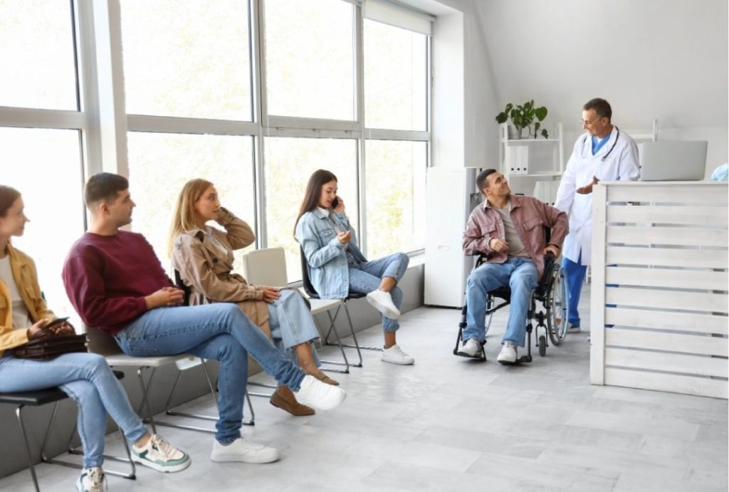 Patients wait for a walk in x ray houston in a bright clinic waiting room as a doctor speaks with a man in a wheelchair.
