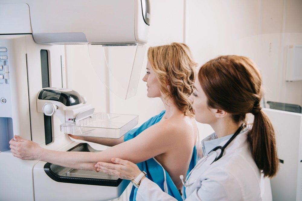 A doctor in a white coat positions a smiling woman at a medical scanning station for mammogram services Houston.