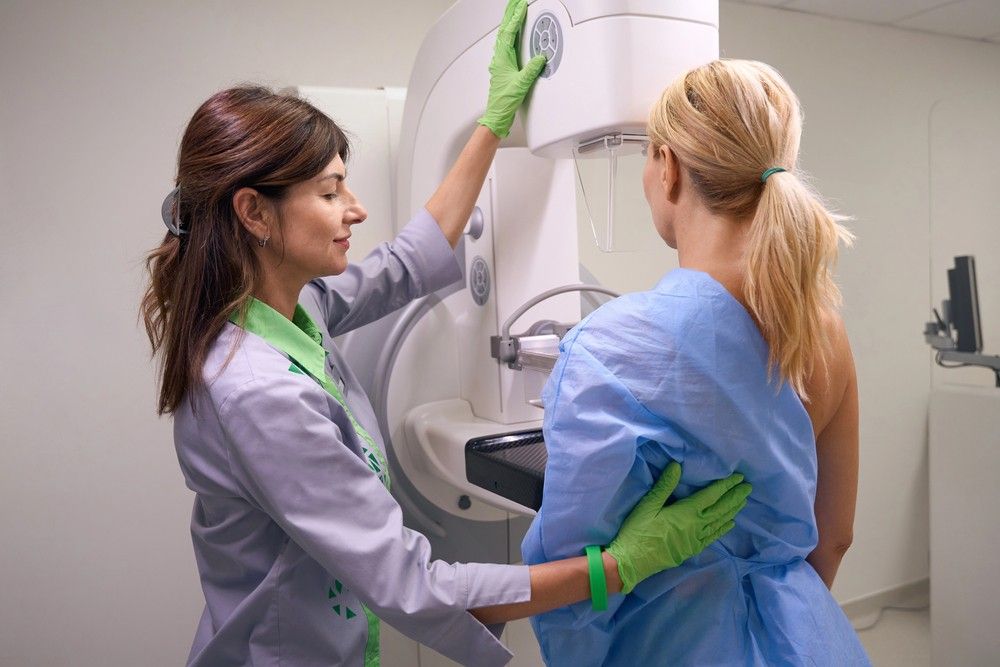 A female technician in green gloves helps a patient use a modern screening machine for mammogram services Houston.